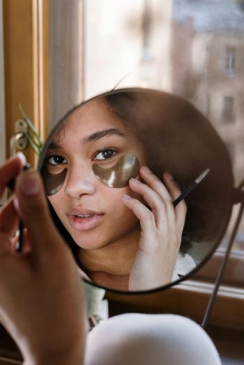 A woman with curly hair applies eye patches in front of a mirror, focusing on skincare.