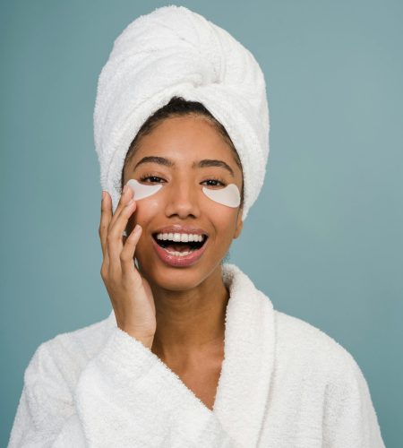 Cheerful young black female in white bathrobe with towel on head and eye patches laughing and looking at camera during skin care routine after shower